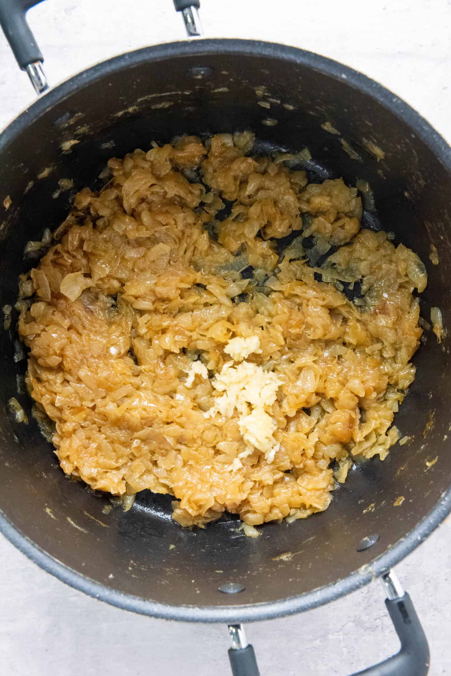Caramelized onions with minced garlic in a black pot, viewed from above on a light-colored surface—perfect beginnings for classic French Onion Soup.
