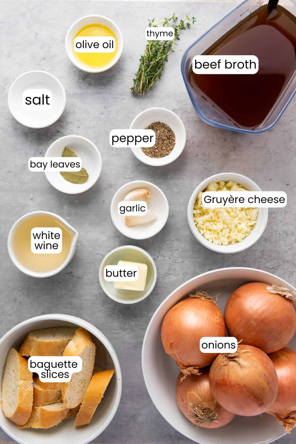Bowls and containers with labeled ingredients for French Onion Soup, including onions, beef broth, Gruyère cheese, baguette slices, butter, garlic, white wine, bay leaves, salt, pepper, olive oil, and thyme.
