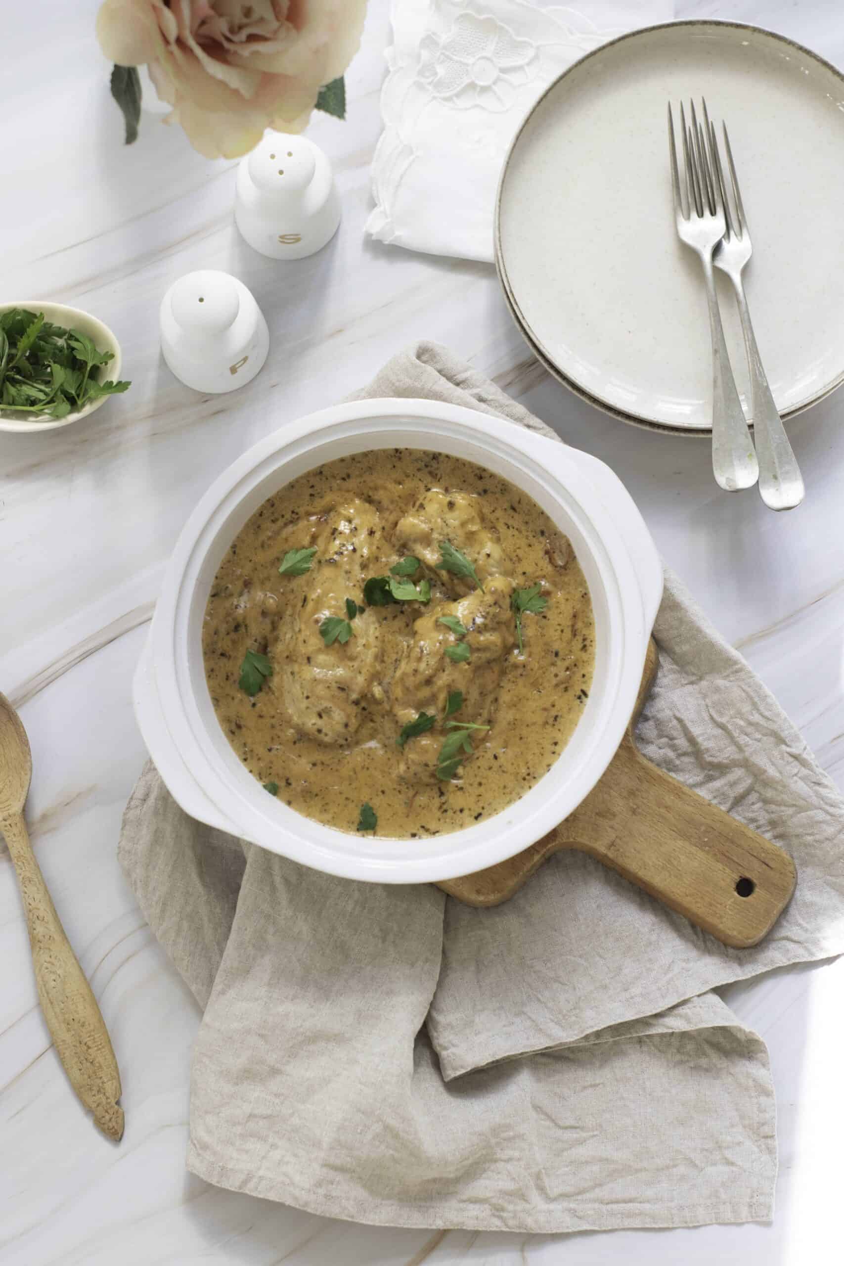 A bowl of creamy chicken curry garnished with herbs, perfect for weeknight dinners, placed on a wooden board with a linen napkin, next to a plate with forks and a flower.