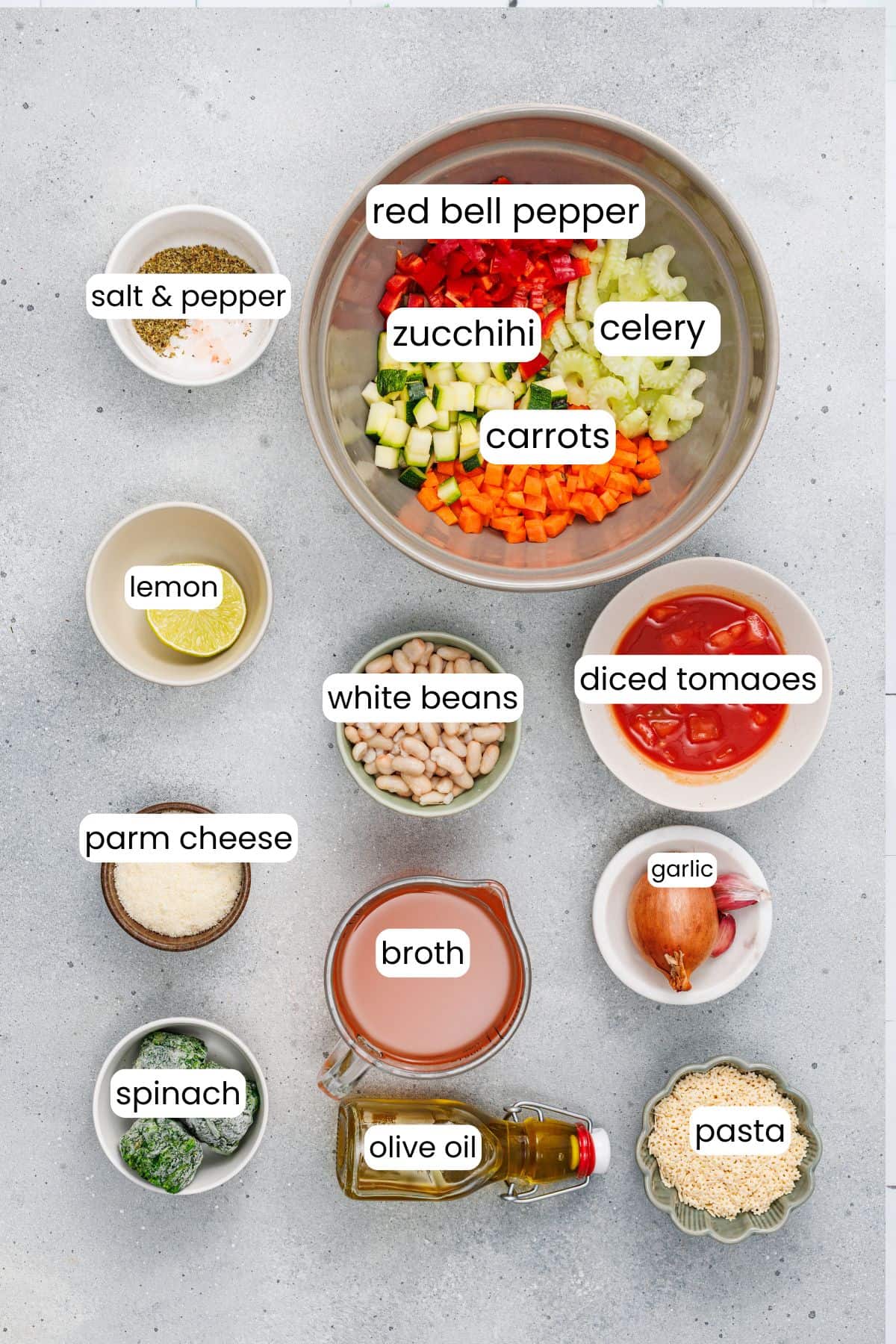 Top-down view of ingredients for a Veggie Minestrone Soup, including chopped vegetables, pasta, broth, olive oil, spinach, parmesan cheese, diced tomatoes, white beans, lemon, and seasonings—perfect for a hearty soup or one-pot dinner.