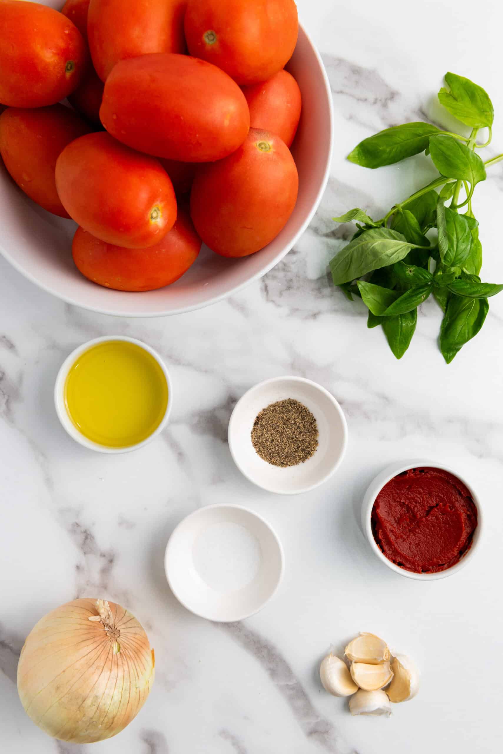 A bowl of Roma tomatoes, fresh basil, olive oil, black pepper, tomato paste, onion, garlic cloves, and salt are arranged on a marble surface—perfect ingredients for a classic Tomato Basil Soup.
