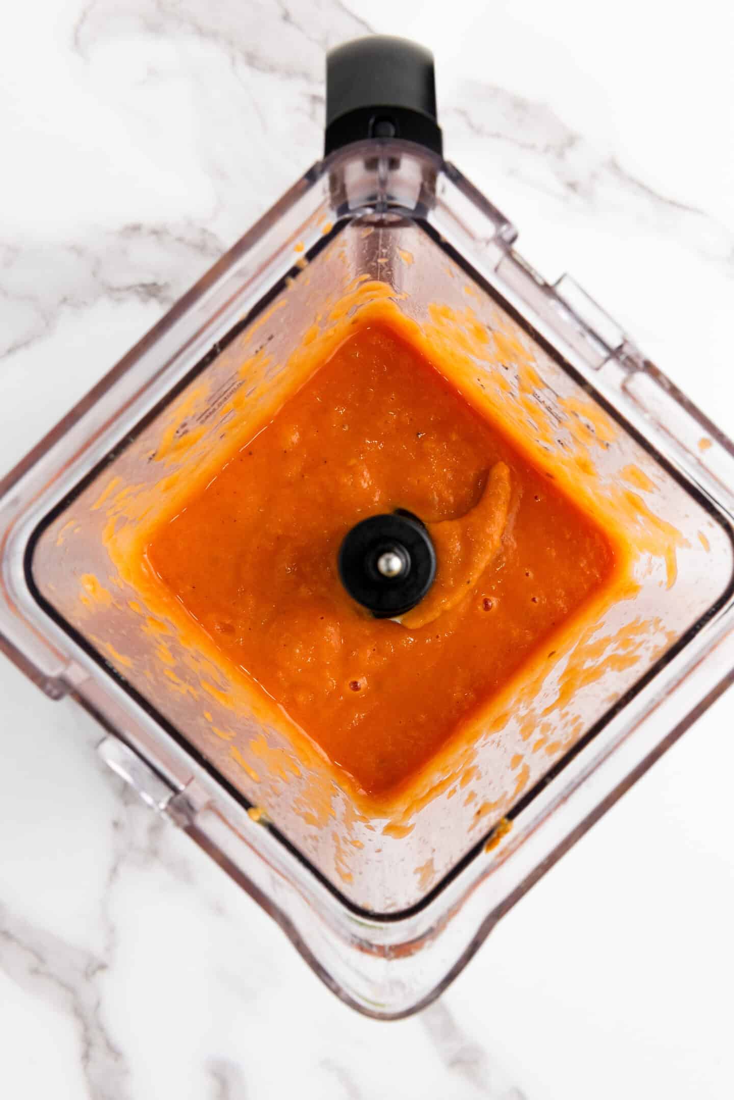 Overhead view of an orange sauce or puree in a blender on a white marble surface, perfect as a base for Roasted Tomato Soup.