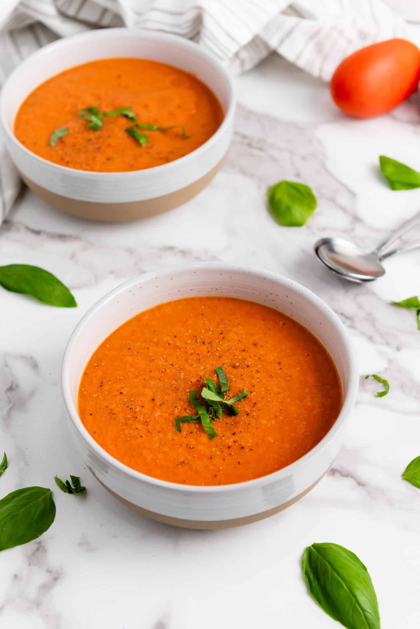 Two bowls of Tomato Basil Soup garnished with fresh herbs rest on a marble surface, surrounded by basil leaves, a spoon, and a striped cloth napkin.