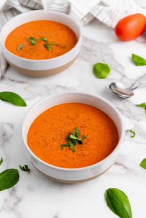 Two bowls of Tomato Basil Soup garnished with fresh herbs rest on a marble surface, surrounded by basil leaves, a spoon, and a striped cloth napkin.