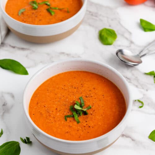 Two bowls of Tomato Basil Soup garnished with fresh herbs rest on a marble surface, surrounded by basil leaves, a spoon, and a striped cloth napkin.