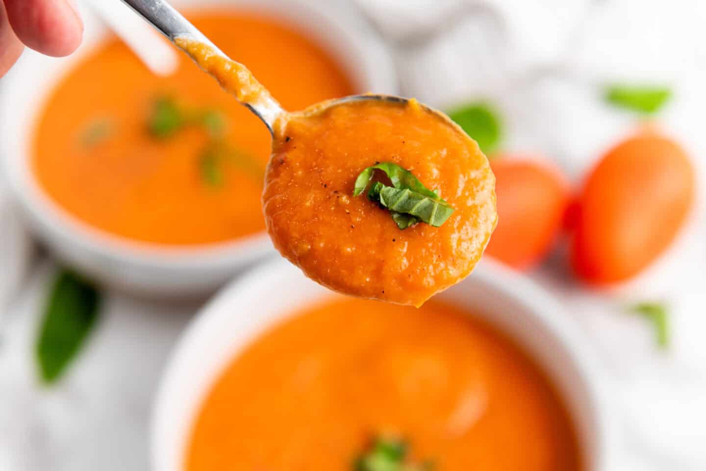 A close-up of a spoonful of creamy roasted tomato soup garnished with fresh basil, with bowls of soup and ripe tomatoes in the background.