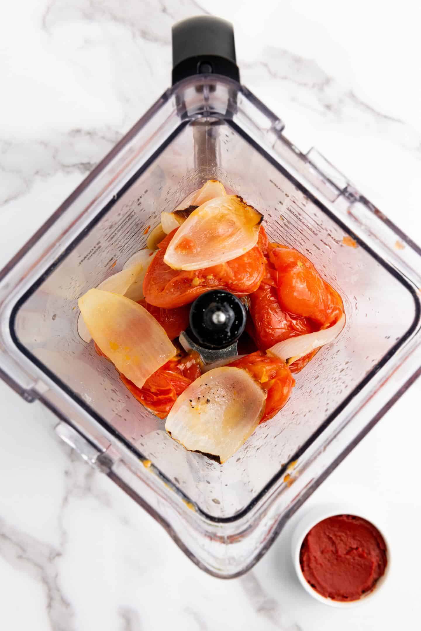 Chopped tomatoes and onion pieces in a blender, viewed from above, ready to create a flavorful Tomato Basil Soup, with a small bowl of tomato paste beside it on a white marble surface.