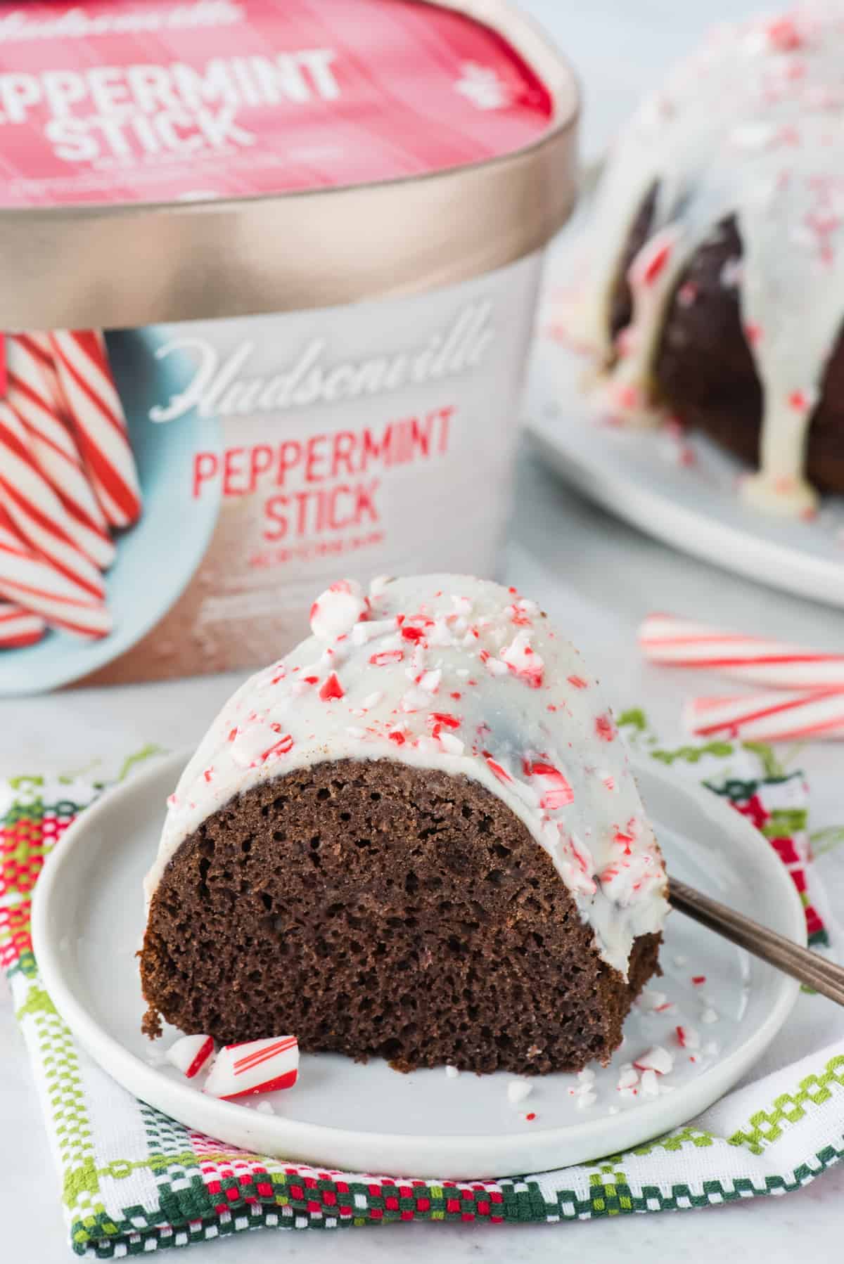 A slice of chocolate bundt cake with white icing and crushed peppermint candy, on a plate with a fork, in front of a tub of Hudsonville Peppermint Stick ice cream—perfect for your Christmas Dinner Menu.
