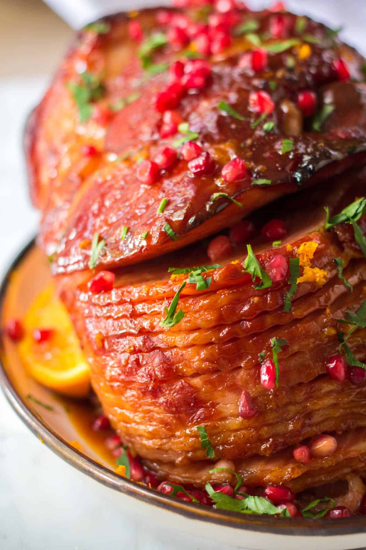 A glazed, sliced ham garnished with chopped herbs and pomegranate seeds sits on a plate with orange slices—an elegant centerpiece for your Christmas Dinner Menu.