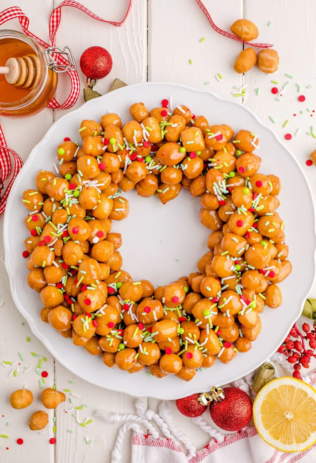 A wreath-shaped arrangement of honey-glazed Italian struffoli, decorated with colorful sprinkles, makes a delightful addition to any Christmas Dinner Menu, presented on a white plate with festive holiday decorations nearby.