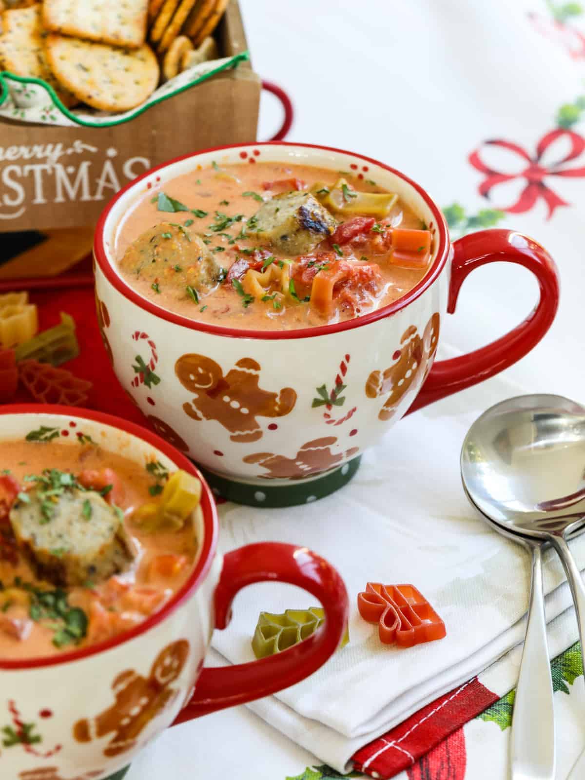 A festive mug filled with creamy tomato soup and pasta shapes, topped with herbs and croutons, sits next to a spoon and Christmas-themed napkin—an inviting starter for your Christmas Dinner Menu, with crackers in the background.