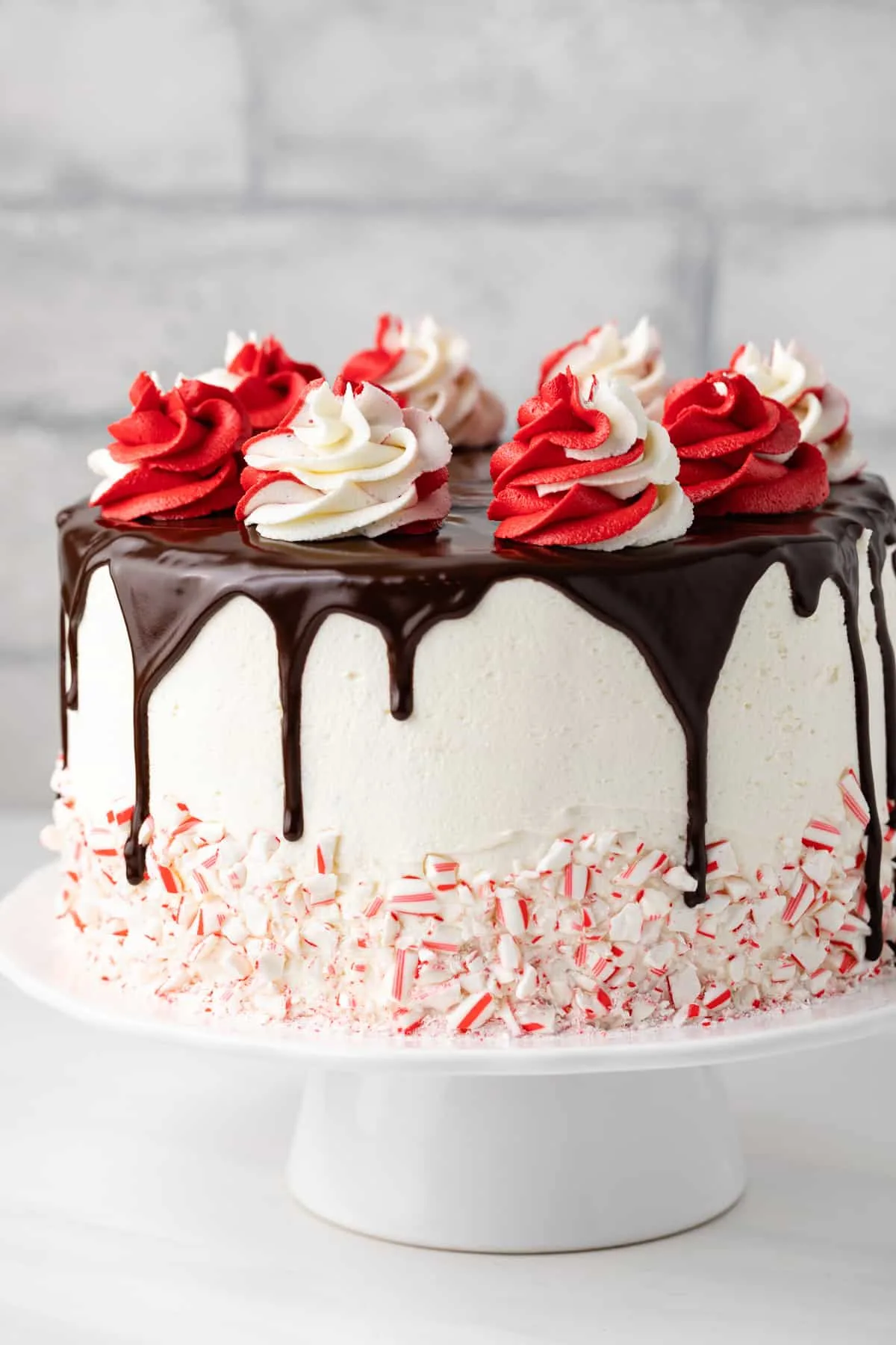 A white frosted cake with chocolate ganache drip, topped with red and white swirled whipped cream and crushed peppermint candies around the base, makes a festive centerpiece for any Christmas Dinner Menu, displayed elegantly on a white cake stand.