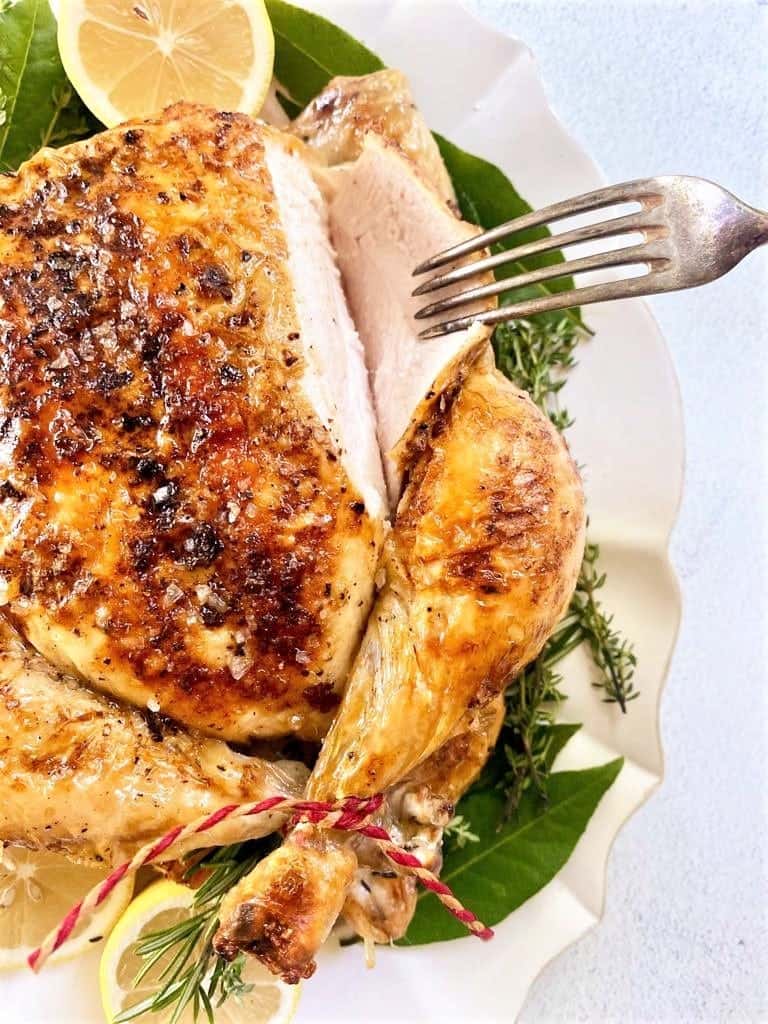 Roast chicken with golden, crispy skin is being sliced on a white plate, garnished with lemon slices and fresh herbs—a perfect addition to your Christmas Dinner Menu. A fork pierces the tender meat, ready to serve.