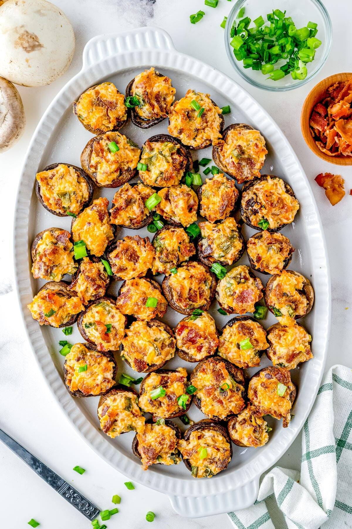 A white oval dish filled with stuffed mushrooms topped with melted cheese and green onions, perfect for your Christmas Dinner Menu, with bowls of bacon bits and chopped green onions nearby.
