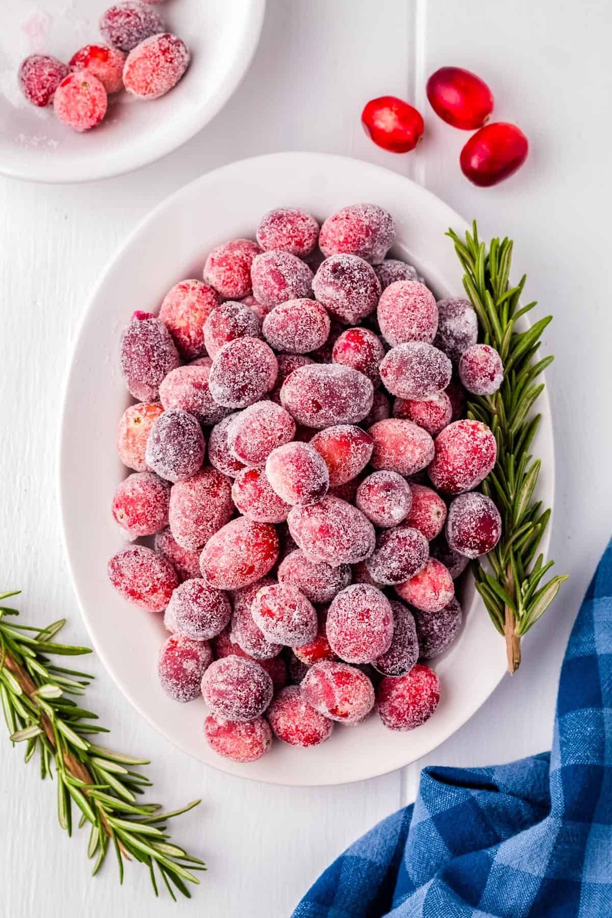 A white plate filled with sugared cranberries, garnished with rosemary sprigs, sits on a white surface next to a blue checkered cloth—perfect for your Christmas Dinner Menu.