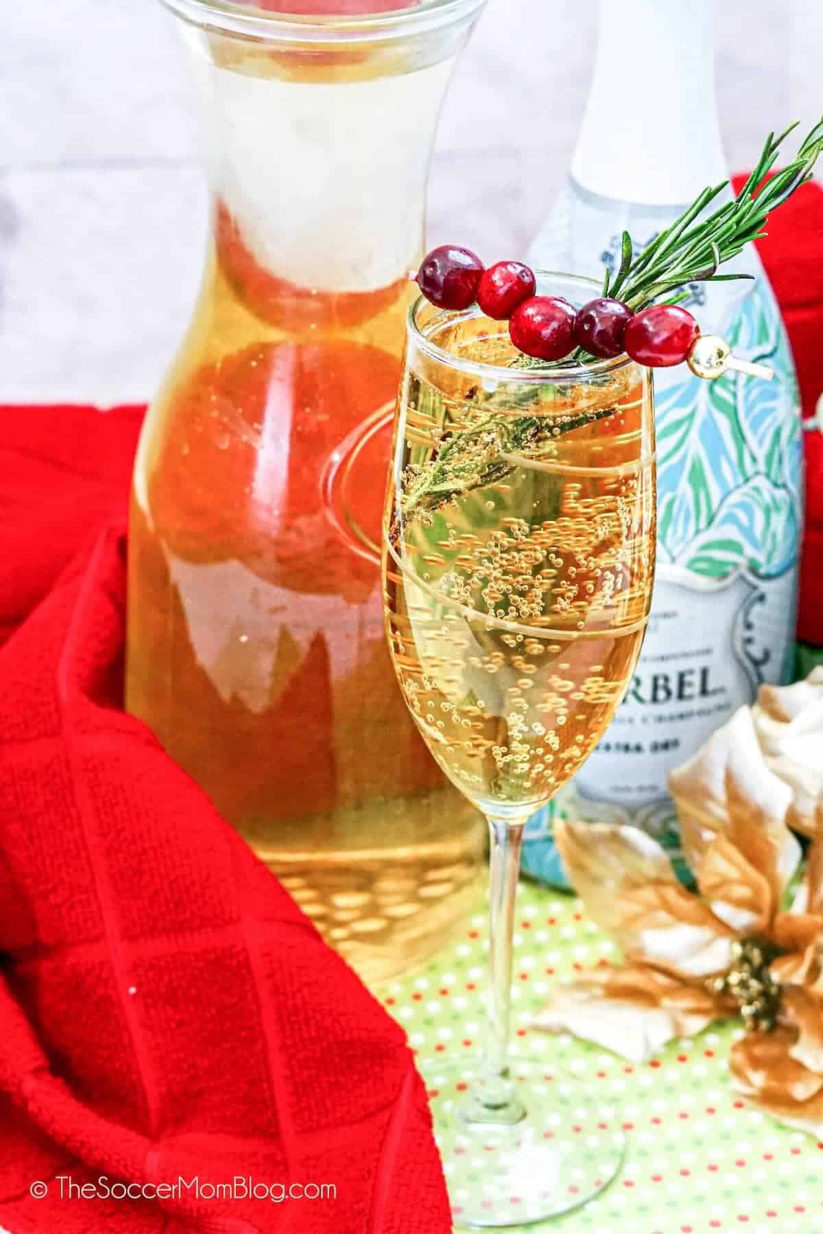 A champagne flute with a cranberry and rosemary garnish, filled with a bubbly drink, sits in front of a glass pitcher, a bottle, and festive red and gold decorations—perfect for elevating your Christmas Dinner Menu.