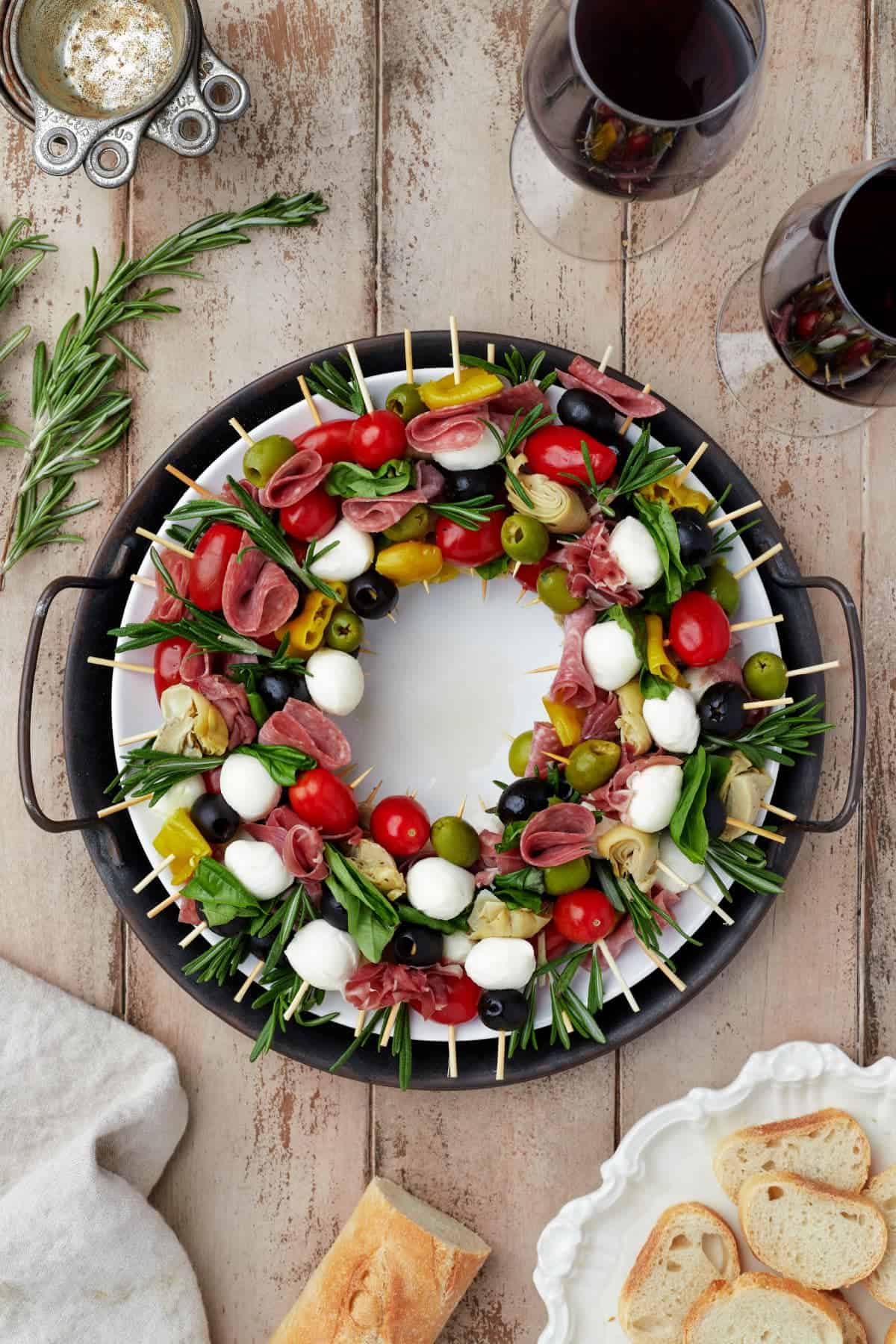 A festive wreath-shaped antipasto platter, perfect for your Christmas Dinner, with skewers of mozzarella, tomatoes, olives, meats, and herbs on a round white plate, surrounded by bread and two glasses of red wine.