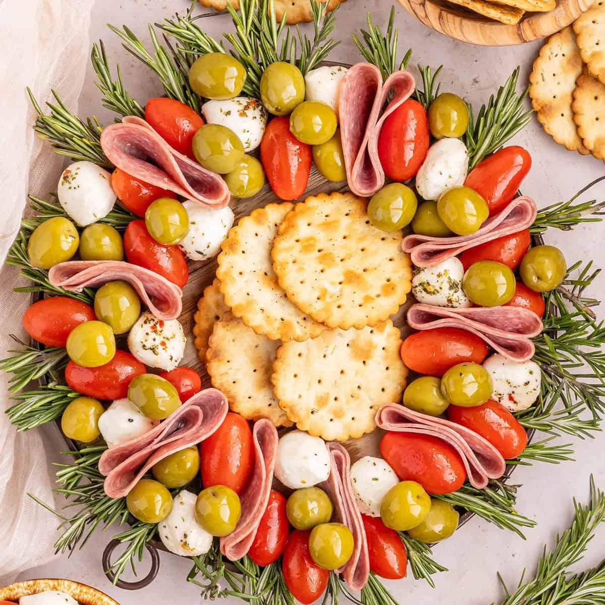 A round platter arranged with crackers, mozzarella balls, green olives, grape tomatoes, folded salami slices, and sprigs of rosemary forms a festive wreath—an eye-catching addition to your Christmas Dinner Menu.