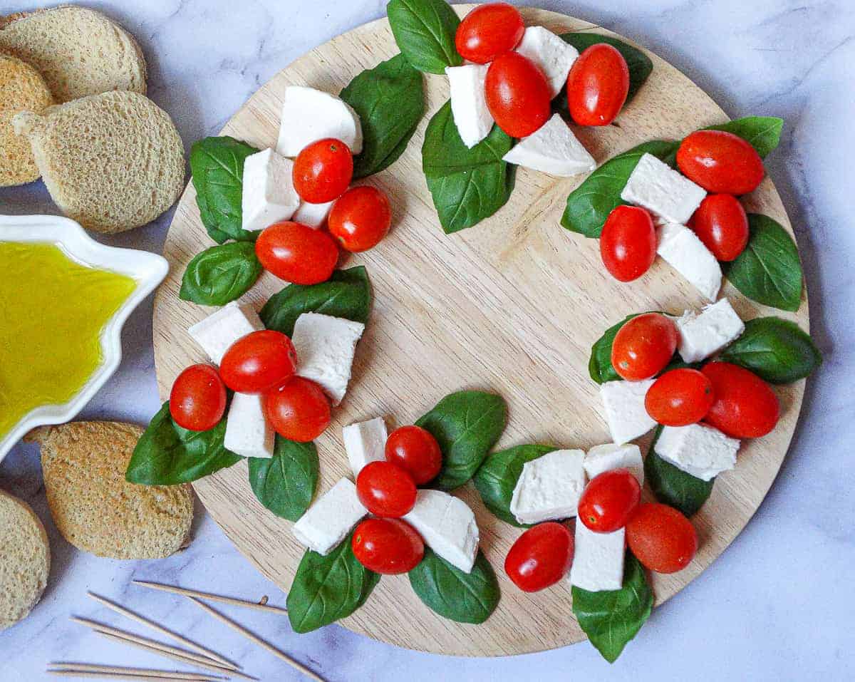 A round wooden board arranged with clusters of basil leaves, mozzarella slices, and cherry tomatoes in a wreath shape, perfect as a festive appetizer for your Christmas Dinner Menu, with bread, olive oil, and toothpicks nearby.