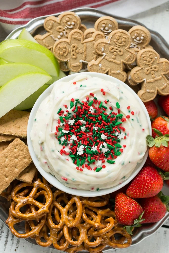 A festive platter for your Christmas Dinner Menu features a bowl of white dip topped with red and green sprinkles, surrounded by gingerbread cookies, apple slices, graham crackers, pretzels, and strawberries.