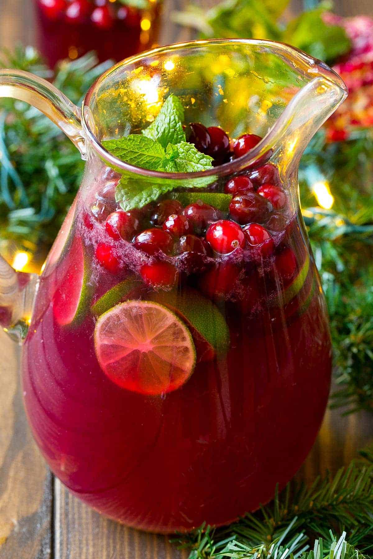 A festive glass pitcher filled with a red beverage garnished with lime slices, cranberries, and mint leaves—perfect for your Christmas Dinner Menu—surrounded by greenery and twinkling string lights.