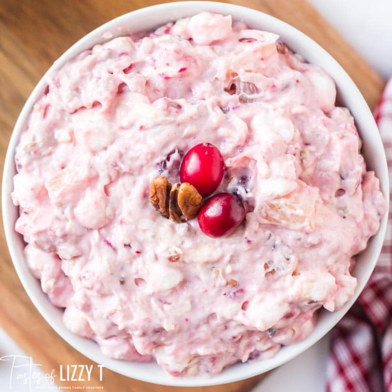 A bowl of creamy pink fruit salad topped with two cranberries and a pecan half, perfect for your Christmas Dinner Menu, placed on a wooden surface.