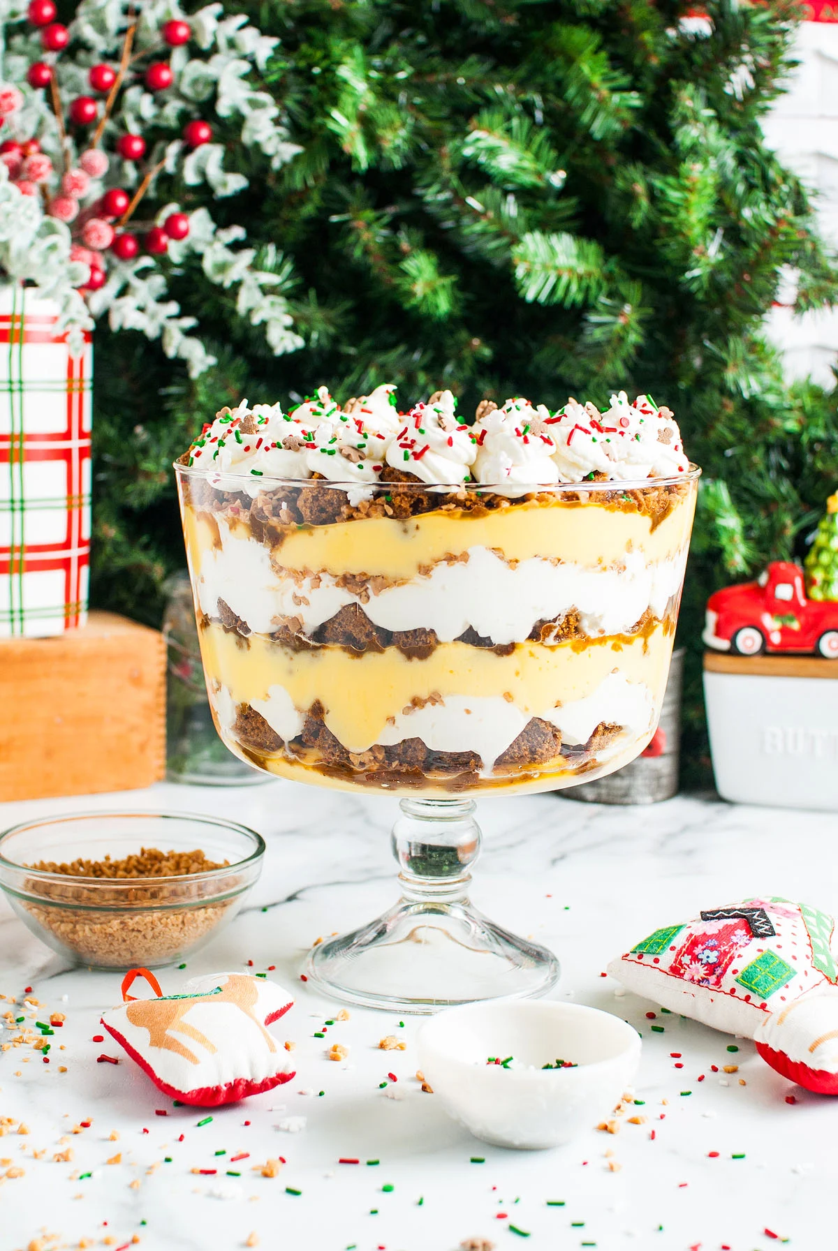 A layered trifle dessert with whipped cream, pudding, and cake in a glass bowl, topped with sprinkles, sits on a table with holiday decorations and a Christmas tree—perfect for your Christmas Dinner Menu.