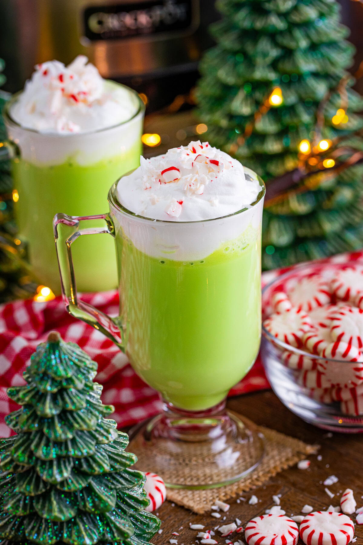 A glass mug with green peppermint drink topped with whipped cream and crushed peppermint candy, set among decorative Christmas trees and a bowl of peppermint candies—perfect for any festive Christmas Dinner or holiday menu.
