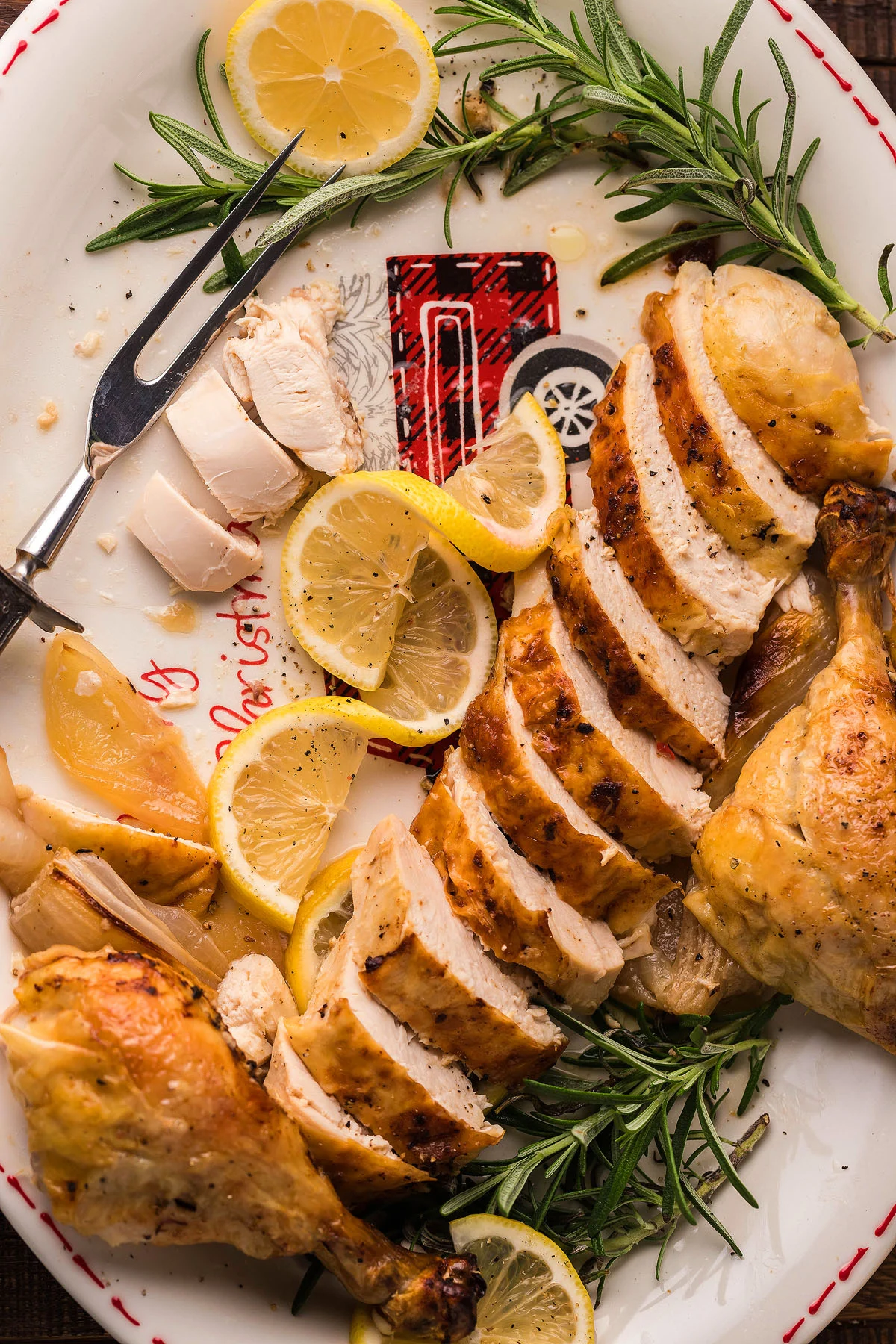 Sliced roasted chicken breast and drumsticks, garnished with lemon slices and fresh rosemary, make a delicious addition to your Christmas Dinner Menu on a white platter.