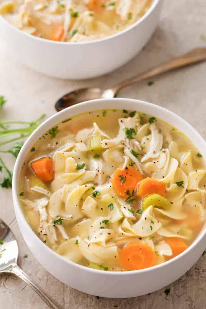 A white bowl of high protein chicken noodle soup with carrots, celery, and parsley garnish sits on a table next to a spoon—perfect for those seeking delicious high protein recipes.
