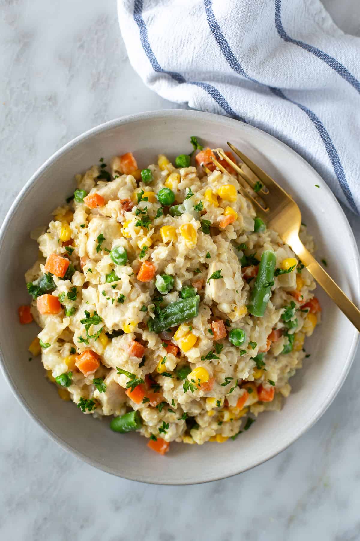 A bowl of mixed vegetable rice with carrots, peas, green beans, and corn—perfect for High Protein recipes—garnished with herbs, beside a gold fork and a striped kitchen towel on a marble surface.
