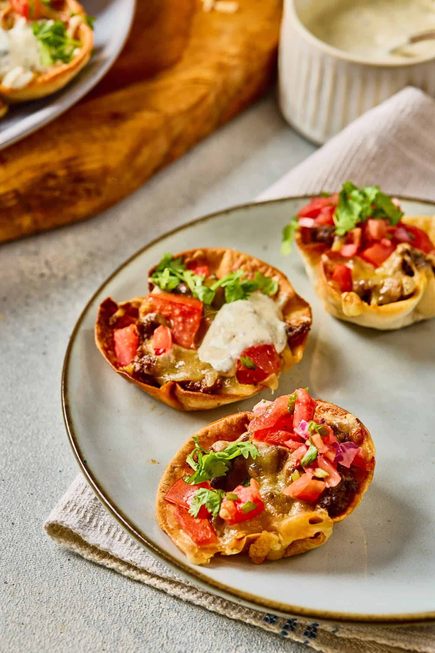 Three mini taco bites filled with beans, cheese, tomatoes, and fresh cilantro are arranged on a plate, with a drizzle of creamy ranch sauce on one and more sauce in a bowl in the background.