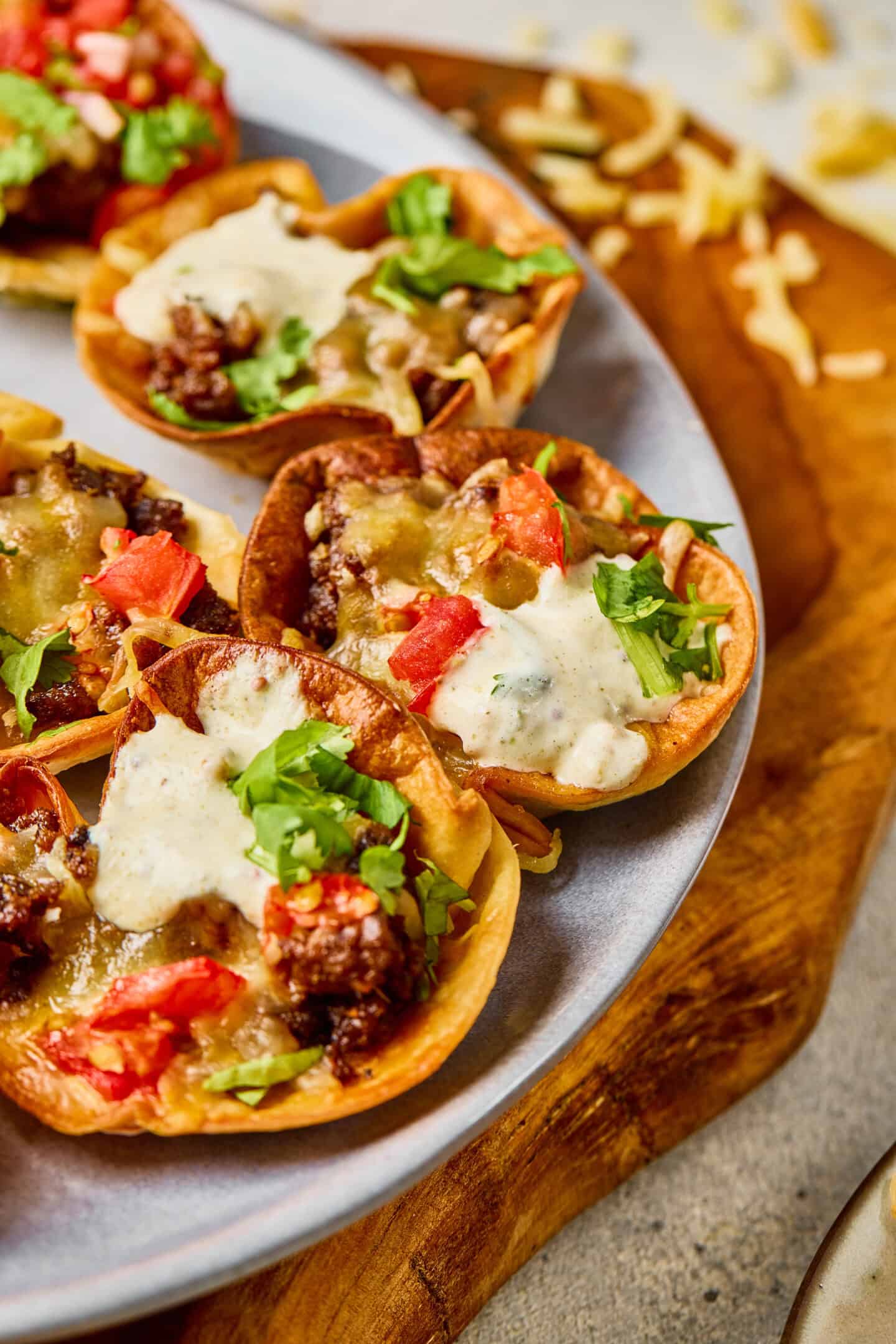 Several mini taco cups filled with ground beef, melted cheese, diced tomatoes, cilantro, and a creamy ranch sauce are arranged on a plate—perfect for savory bites at any gathering.