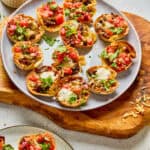 A plate of mini taco cups bites filled with ground meat, cheese, tomatoes, and cilantro, served with two dipping sauces, including ranch, on the side.