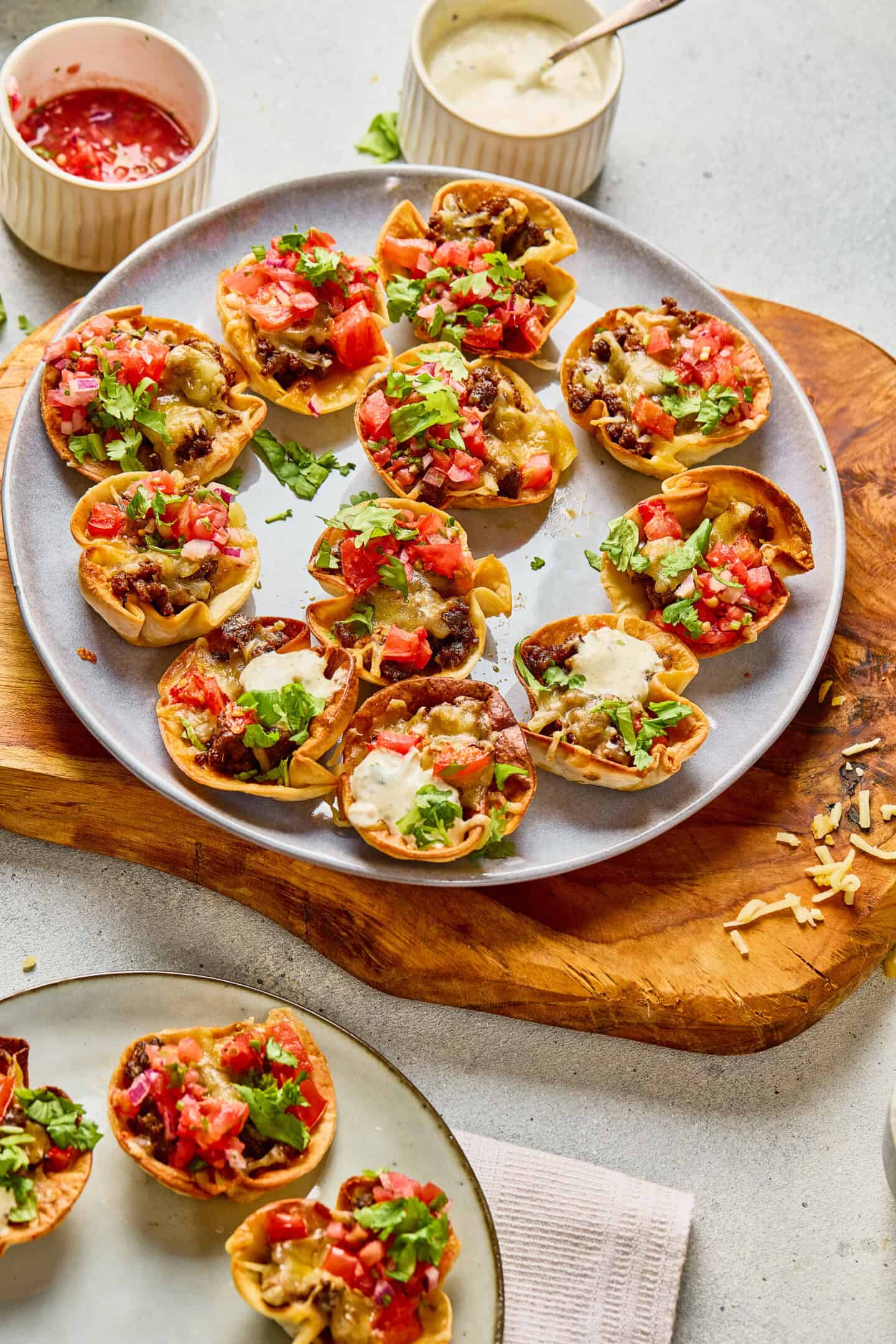 A plate of mini taco bites filled with ground meat, cheese, chopped tomatoes, and cilantro, served on a wooden board with two dipping sauces—including creamy ranch—in small bowls nearby.