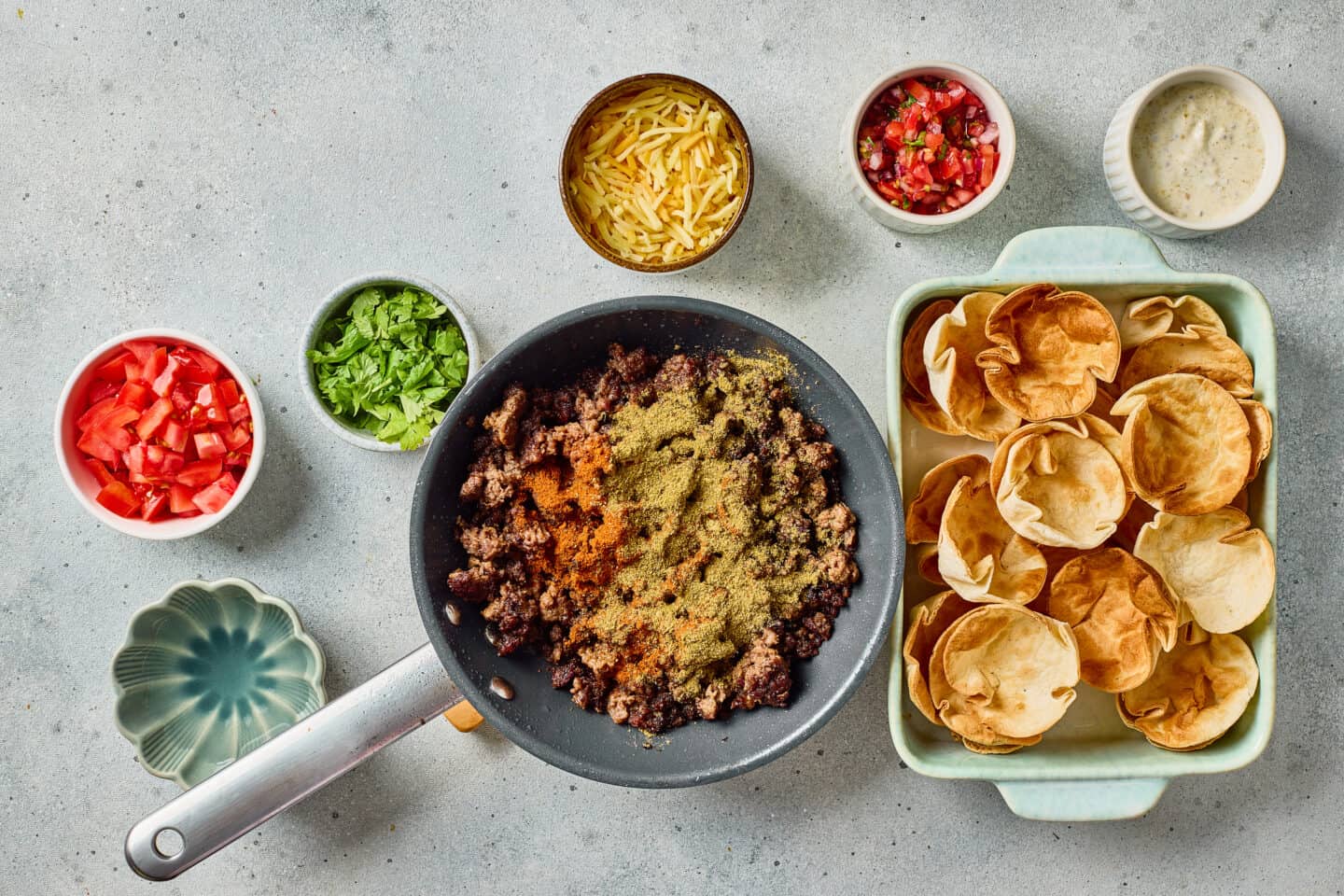 A skillet of seasoned ground beef surrounded by bowls of shredded cheese, chopped lettuce, diced tomatoes, salsa, ranch sauce, and a dish of crispy taco bites.