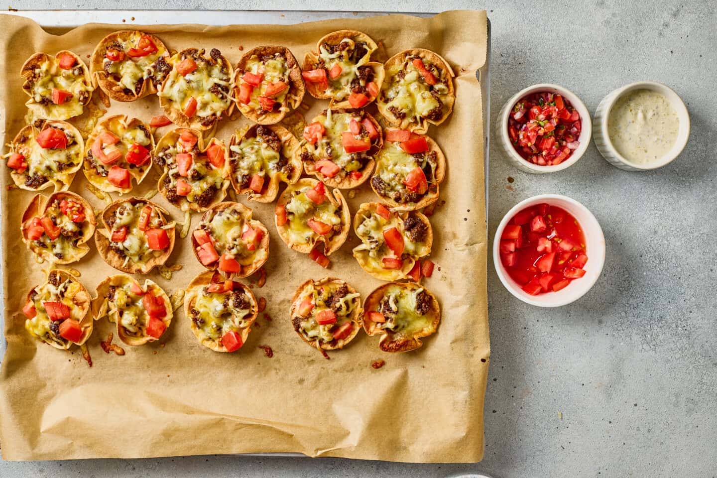 Mini nacho bites topped with melted cheese, ground meat, and diced tomatoes on a parchment-lined baking sheet, with sides of salsa, ranch sauce, and chopped tomatoes in small bowls.