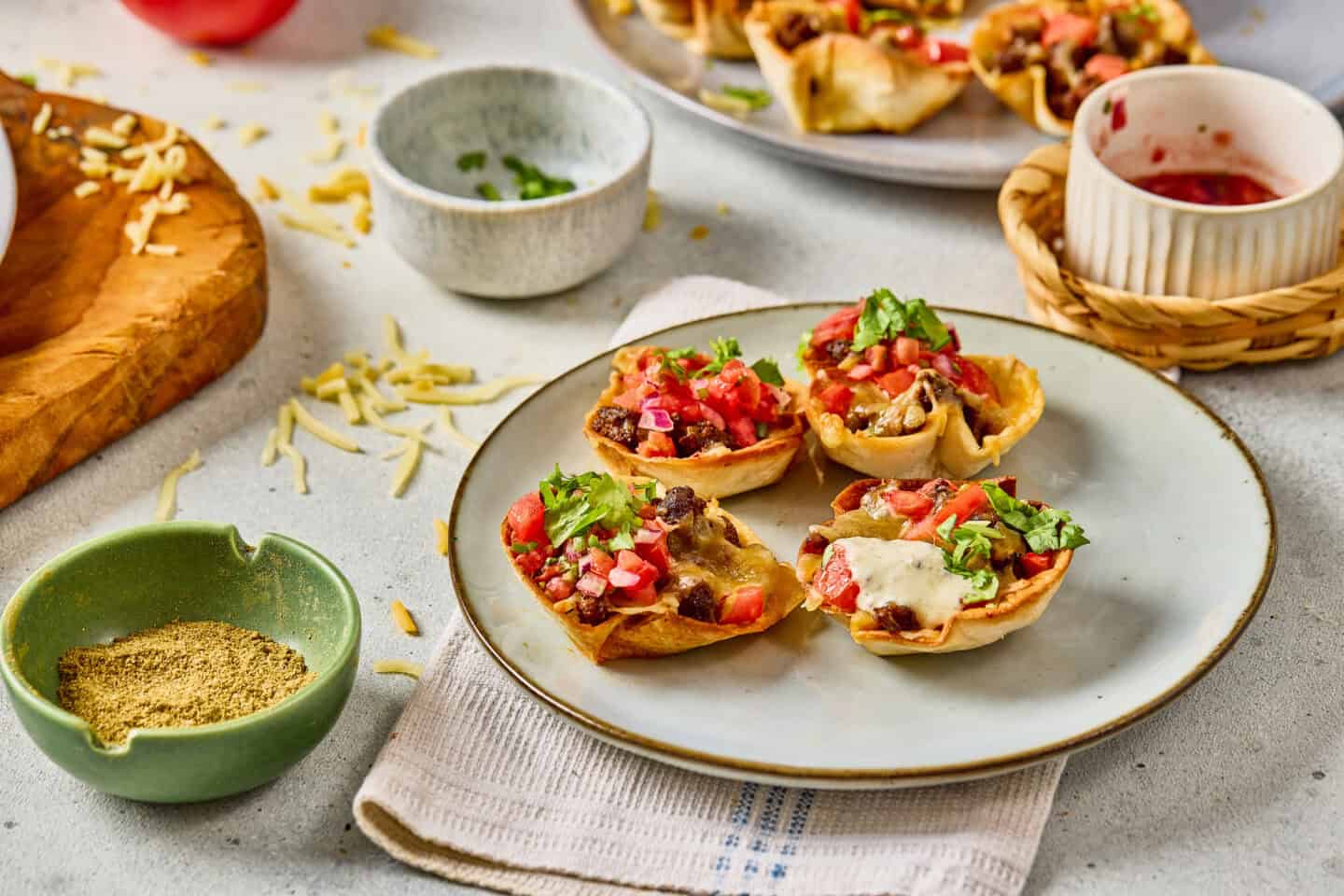 A plate with four mini nacho cups—perfect taco bites—filled with beans, cheese, salsa, and cilantro, surrounded by bowls of shredded cheese, seasoning, and salsa on a light countertop.