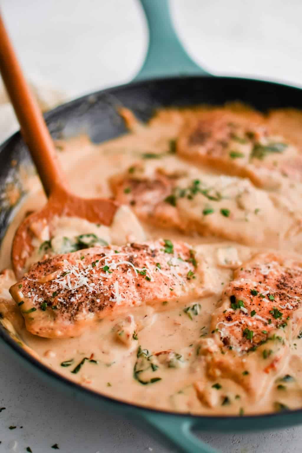 Chicken breasts simmering in a creamy sauce with visible herbs and grated cheese, cooked in a green-handled skillet and stirred with a wooden spoon—perfect for easy family dinner or Sunday dinner ideas.