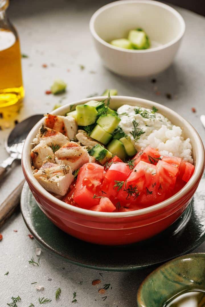 A Mediterranean Chicken Bowl featuring grilled lemon-dill chicken, diced tomatoes, cucumber, white rice, and a dollop of sauce, garnished with fresh dill. In the background are a bowl of cucumbers and oil.