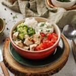A Mediterranean bowl with rice, diced tofu, chopped cucumber, tomato, and a creamy topping, garnished with dill—set on a wooden board with utensils and other Chicken Bowl dishes nearby.