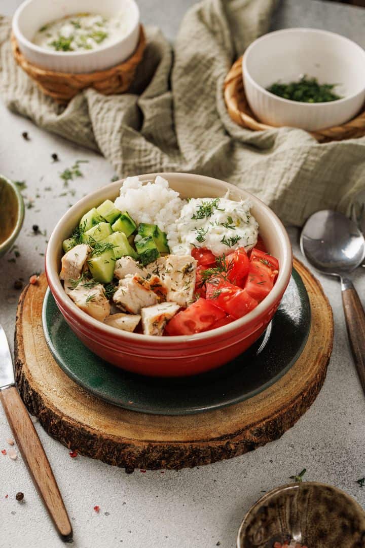 A Mediterranean bowl with rice, diced chicken, chopped cucumber, tomato, and a creamy topping, garnished with dill—set on a wooden board with utensils and other Chicken Bowl dishes nearby.