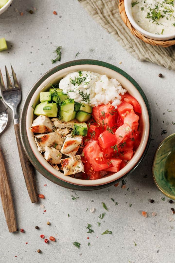 A Mediterranean bowl featuring lemon dill chicken, diced cucumbers, tomatoes, white rice, and creamy sauce, all garnished with fresh dill. A fork and knife are placed beside the chicken bowl for a perfect meal experience.