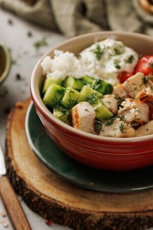 A Mediterranean Chicken Bowl featuring sliced Lemon-Dill Chicken, cucumber, tomato, white rice, and a dollop of yogurt garnished with fresh dill.