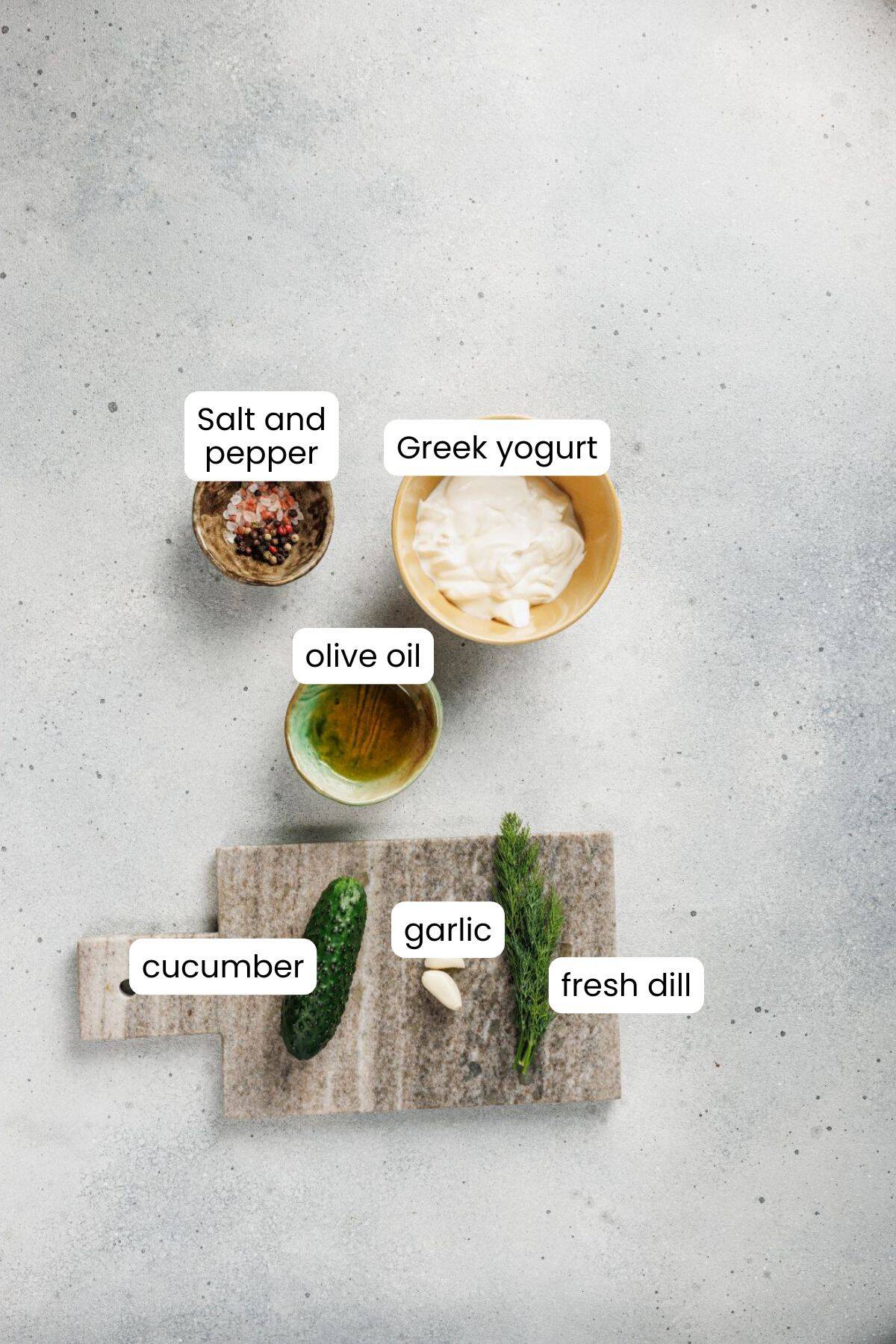 Top view of labeled ingredients for a Mediterranean recipe: salt and pepper, Greek yogurt, olive oil, cucumber, garlic, and fresh dill on a light surface with a small cutting board—perfect for a Lemon-Dill Chicken Bowl.