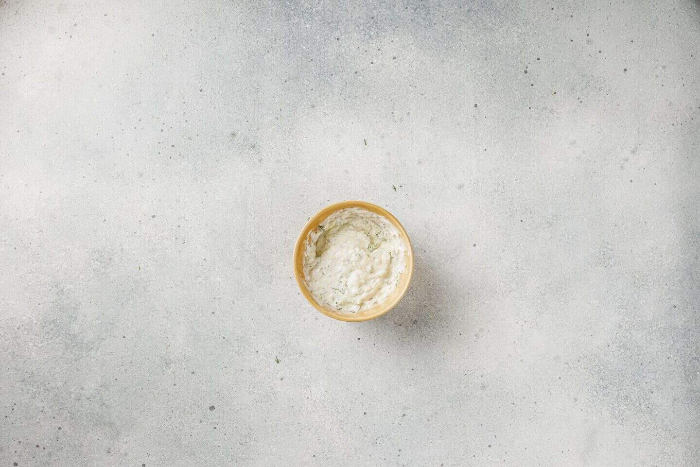A small bowl filled with a creamy white Mediterranean dip is placed on a light gray textured surface.