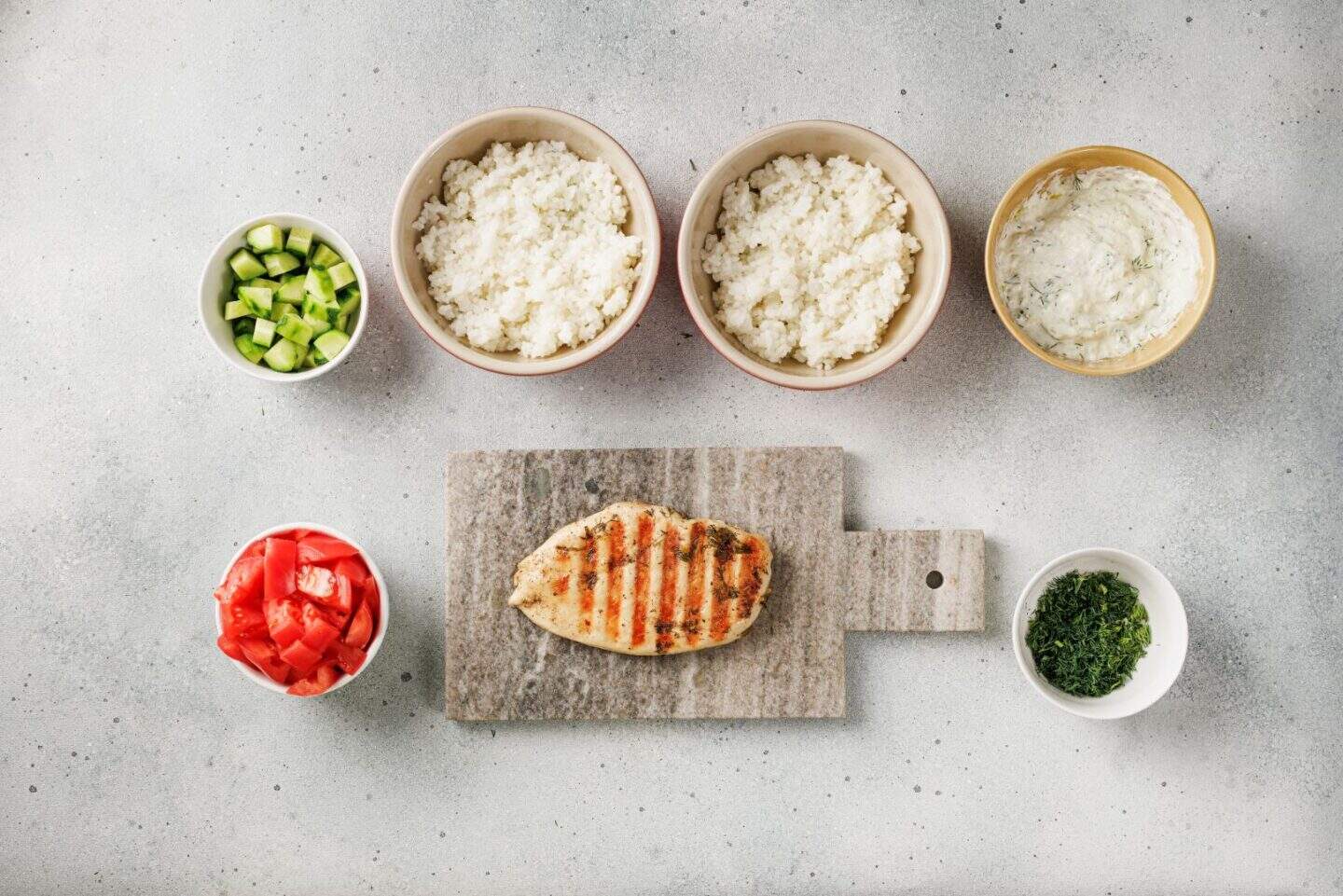 Mediterranean Lemon-Dill Chicken breast on a cutting board, bowls of rice, diced cucumber, tomato, chopped herbs, and a bowl of creamy white sauce on a light surface.