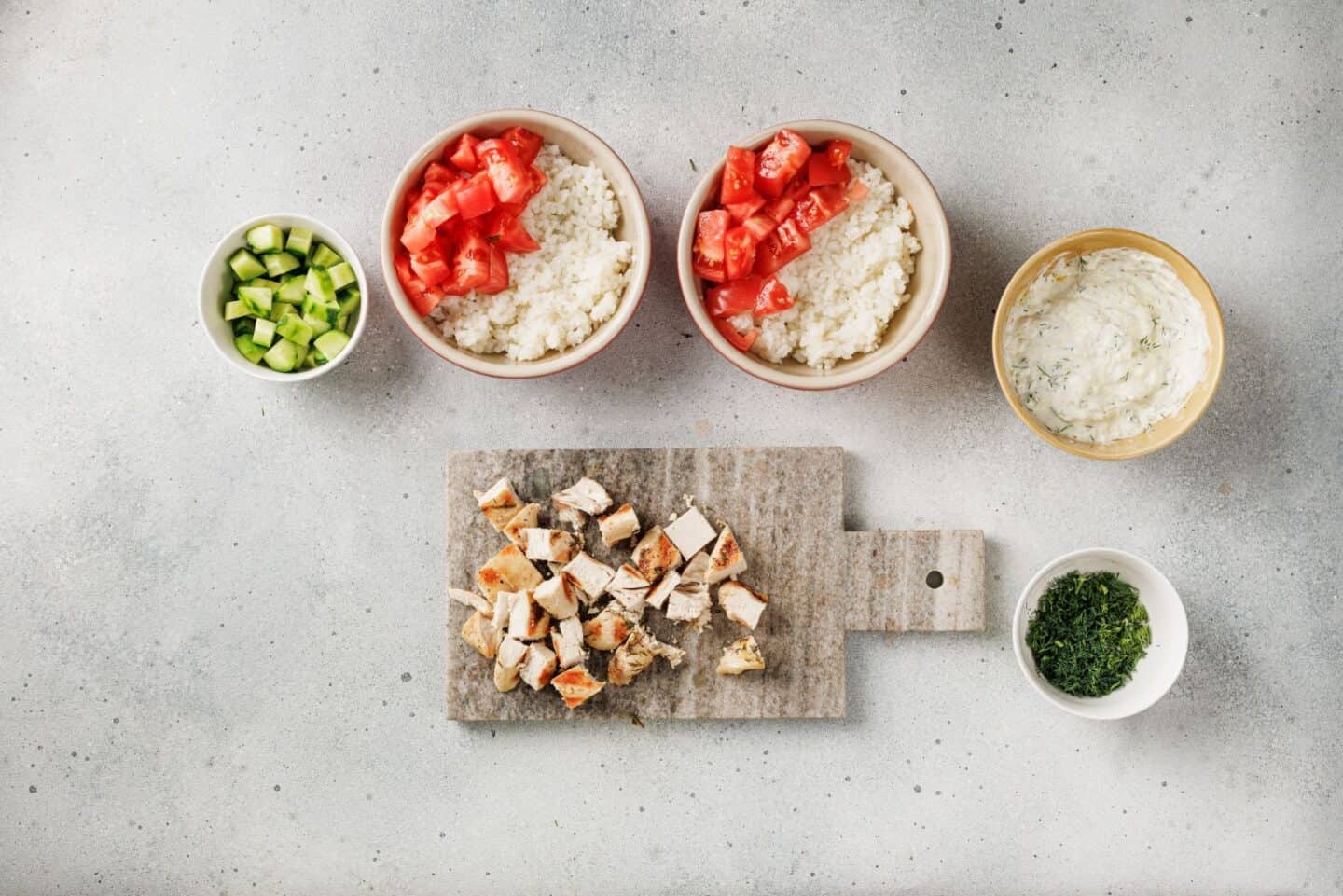 A vibrant Mediterranean spread features two Chicken Bowls with diced tomatoes, a small bowl of chopped cucumber, savory sauce, fresh herbs, and Lemon-Dill Chicken cubes arranged on a cutting board.