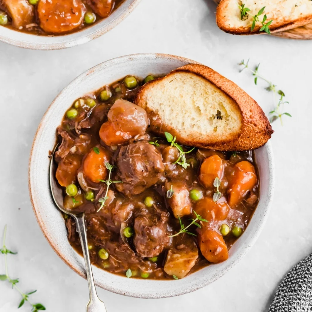 A bowl of high protein beef stew with carrots, peas, and potatoes, garnished with herbs and served with a slice of toasted bread—a comforting choice for Crockpot recipes.