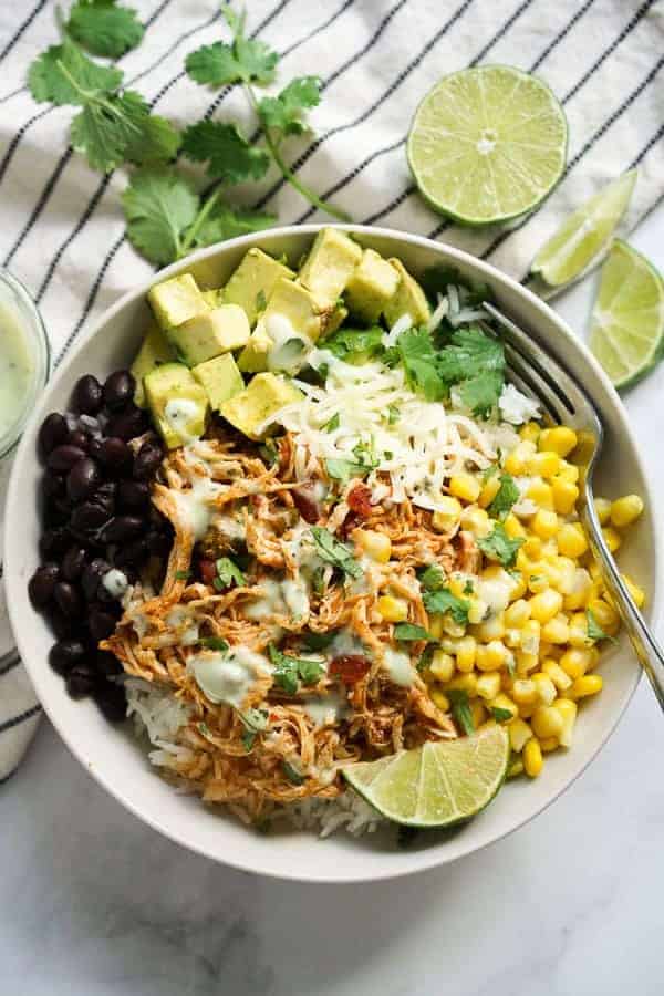 A bowl with rice, shredded chicken, black beans, corn, avocado, cheese, cilantro, and a lime wedge—a delicious high protein recipe perfect for your collection of crockpot recipes. Served on a white cloth with a fork and lime slices nearby.
